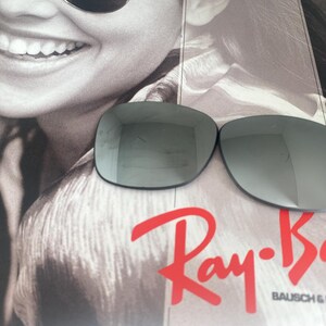 K&ouml;nnte beinhalten: Zwei dunkelgraue Ersatz-Sonnenbrillengl&auml;ser mit reflektierender Oberfl&auml;che. Die Gl&auml;ser liegen auf einem Werbedruck mit einem l&auml;chelnden Gesicht und dem roten Ray-Ban Bausch & Lomb Logo. Die Gl&auml;ser haben eine glatte, geschwungene Form.