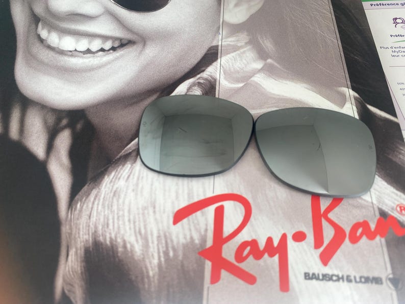 K&ouml;nnte beinhalten: Zwei dunkelgraue Ersatz-Sonnenbrillengl&auml;ser mit reflektierender Oberfl&auml;che. Die Gl&auml;ser liegen auf einem Werbedruck mit einem l&auml;chelnden Gesicht und dem roten Ray-Ban Bausch & Lomb Logo. Die Gl&auml;ser haben eine glatte, geschwungene Form.
