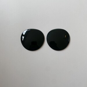 K&ouml;nnte beinhalten: Zwei kreisf&ouml;rmige, dunkel get&ouml;nte Ersatzgl&auml;ser f&uuml;r eine Sonnenbrille. Das linke Glas zeigt das wei&szlig;e Ray-Ban-Logo, das nahe am oberen Rand aufgedruckt ist. Beide Gl&auml;ser haben eine glatte, reflektierende Oberfl&auml;che.