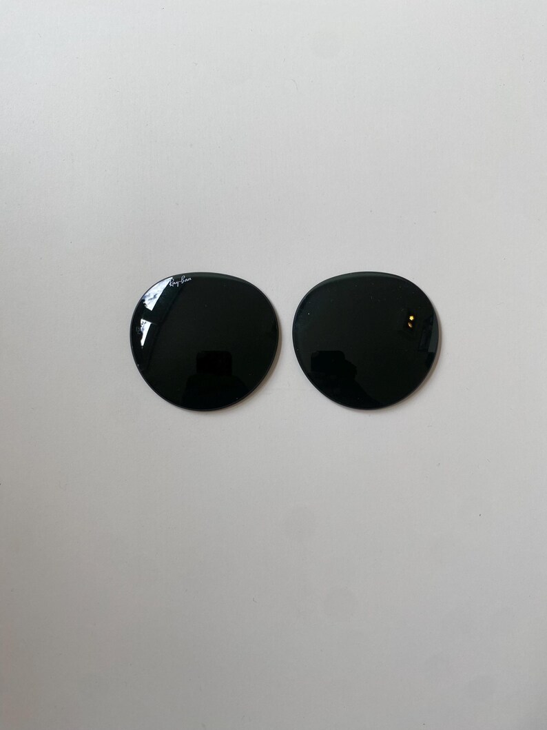 K&ouml;nnte beinhalten: Zwei kreisf&ouml;rmige, dunkel get&ouml;nte Ersatzgl&auml;ser f&uuml;r eine Sonnenbrille. Das linke Glas zeigt das wei&szlig;e Ray-Ban-Logo, das nahe am oberen Rand aufgedruckt ist. Beide Gl&auml;ser haben eine glatte, reflektierende Oberfl&auml;che.