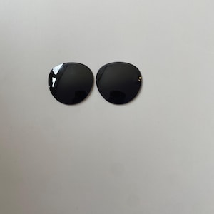 K&ouml;nnte beinhalten: Zwei kreisf&ouml;rmige, tiefschwarze Ersatzgl&auml;ser f&uuml;r eine Sonnenbrille. Die Gl&auml;ser haben eine glatte, reflektierende Oberfl&auml;che mit einer dunklen T&ouml;nung und sind f&uuml;r den Einsatz in Brillenfassungen vorgesehen. Jedes Glas ist kreisrund.