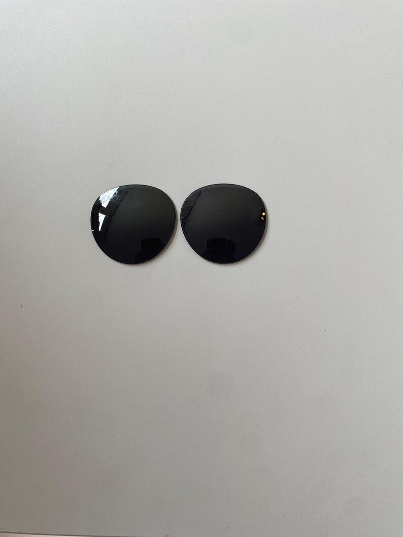 K&ouml;nnte beinhalten: Zwei kreisf&ouml;rmige, tiefschwarze Ersatzgl&auml;ser f&uuml;r eine Sonnenbrille. Die Gl&auml;ser haben eine glatte, reflektierende Oberfl&auml;che mit einer dunklen T&ouml;nung und sind f&uuml;r den Einsatz in Brillenfassungen vorgesehen. Jedes Glas ist kreisrund.