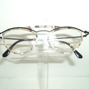 May include: A pair of tortoiseshell spectacles with black metal frames. The glasses have a cat-eye shape and are on a clear display stand.