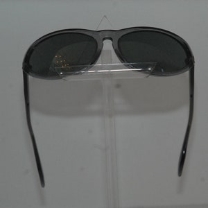 Pode incluir: Um par de &oacute;culos de sol aviador preto com lentes escuras. Os &oacute;culos est&atilde;o em um suporte transparente.