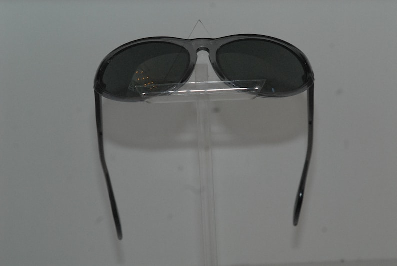 Pode incluir: Um par de &oacute;culos de sol aviador preto com lentes escuras. Os &oacute;culos est&atilde;o em um suporte transparente.