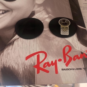 K&ouml;nnte beinhalten: Zwei kreisf&ouml;rmige, dunkelgraue Brillengl&auml;ser auf einer Werbekarte von Ray-Ban Bausch & Lomb. Ein Glas ist mit einem goldenen Aufkleber versehen, der das Ray-Ban-Logo, den Text G-15 LENS und das Symbol der olympischen Ringe zeigt.