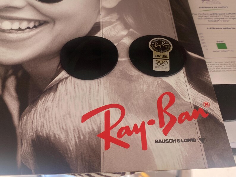 K&ouml;nnte beinhalten: Zwei kreisf&ouml;rmige, dunkelgraue Brillengl&auml;ser auf einer Werbekarte von Ray-Ban Bausch & Lomb. Ein Glas ist mit einem goldenen Aufkleber versehen, der das Ray-Ban-Logo, den Text G-15 LENS und das Symbol der olympischen Ringe zeigt.