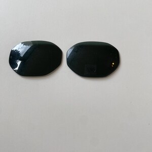 K&ouml;nnte beinhalten: Zwei dunkle, oval geformte Ersatzgl&auml;ser f&uuml;r eine Sonnenbrille mit einer glatten, reflektierenden Oberfl&auml;che. Das rechte Glas weist in der Mitte den kleinen, eingravierten Zahlencode 5395 auf, der zur Identifizierung des Modells dient.