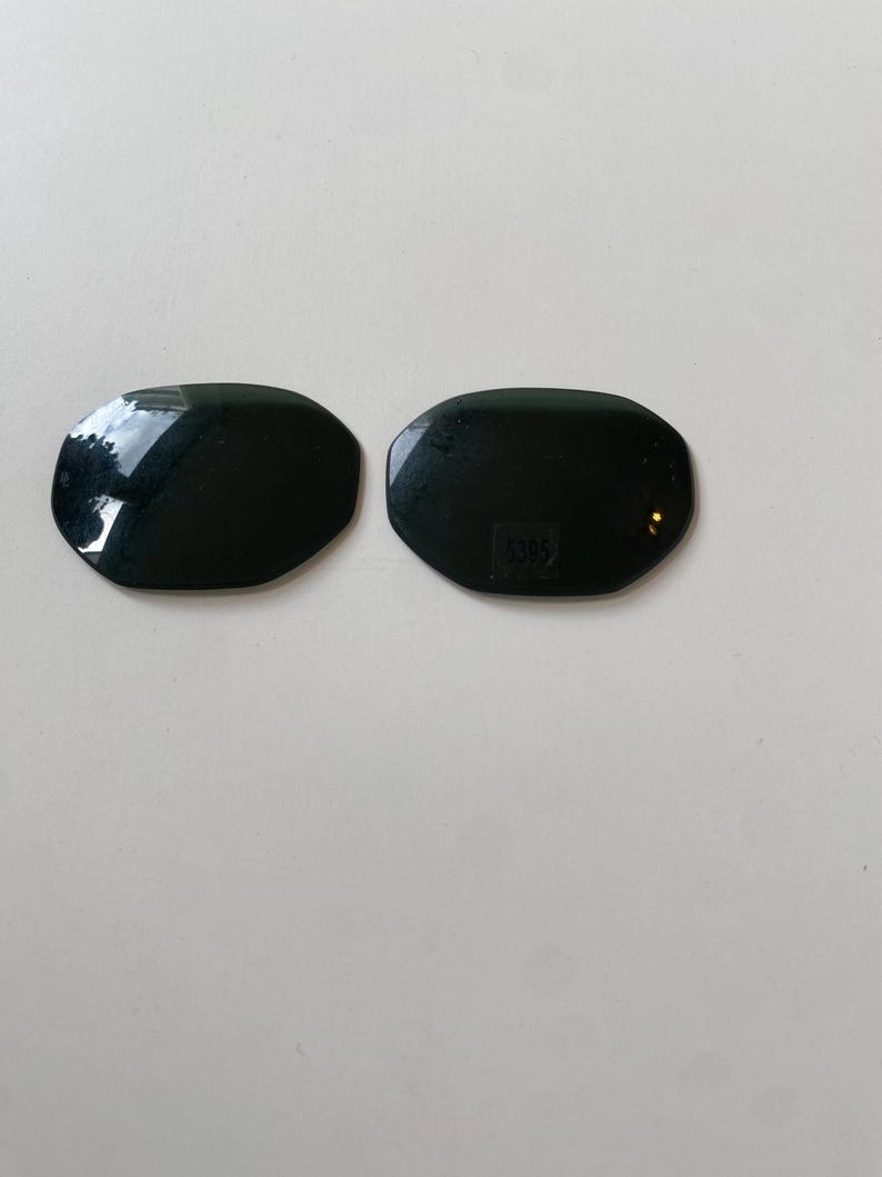 K&ouml;nnte beinhalten: Zwei dunkle, oval geformte Ersatzgl&auml;ser f&uuml;r eine Sonnenbrille mit einer glatten, reflektierenden Oberfl&auml;che. Das rechte Glas weist in der Mitte den kleinen, eingravierten Zahlencode 5395 auf, der zur Identifizierung des Modells dient.