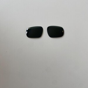 K&ouml;nnte beinhalten: Zwei dunkelgr&uuml;ne Ersatzgl&auml;ser f&uuml;r Sonnenbrillen in rechteckiger Form mit abgerundeten Ecken. Das linke Glas zeigt das wei&szlig;e Ray-Ban-Logo in der oberen Ecke. Diese Gl&auml;ser sind f&uuml;r Brillenrahmen konzipiert und haben eine glatte, reflektierende Oberfl&auml;che.