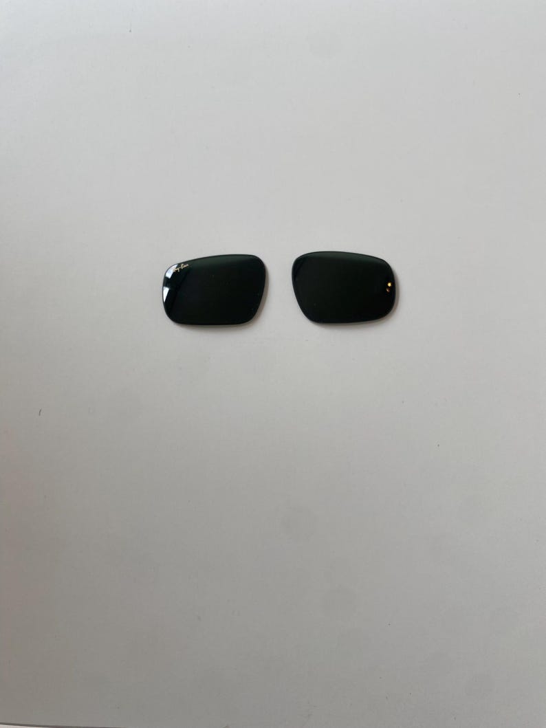 K&ouml;nnte beinhalten: Zwei dunkelgr&uuml;ne Ersatzgl&auml;ser f&uuml;r Sonnenbrillen in rechteckiger Form mit abgerundeten Ecken. Das linke Glas zeigt das wei&szlig;e Ray-Ban-Logo in der oberen Ecke. Diese Gl&auml;ser sind f&uuml;r Brillenrahmen konzipiert und haben eine glatte, reflektierende Oberfl&auml;che.