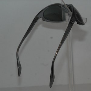 Pode incluir: Um par de &oacute;culos de sol cinzentos com arma&ccedil;&atilde;o curva e lentes cinzas. Os &oacute;culos de sol s&atilde;o exibidos em um suporte transparente.