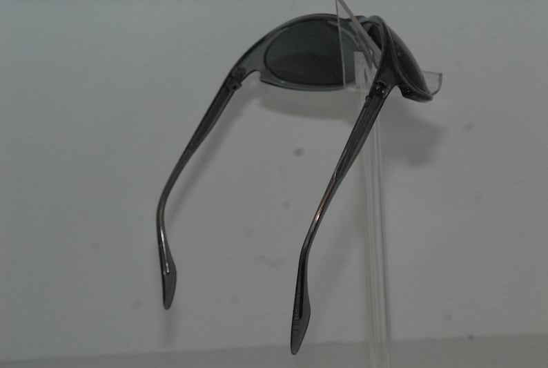 Pode incluir: Um par de &oacute;culos de sol cinzentos com arma&ccedil;&atilde;o curva e lentes cinzas. Os &oacute;culos de sol s&atilde;o exibidos em um suporte transparente.