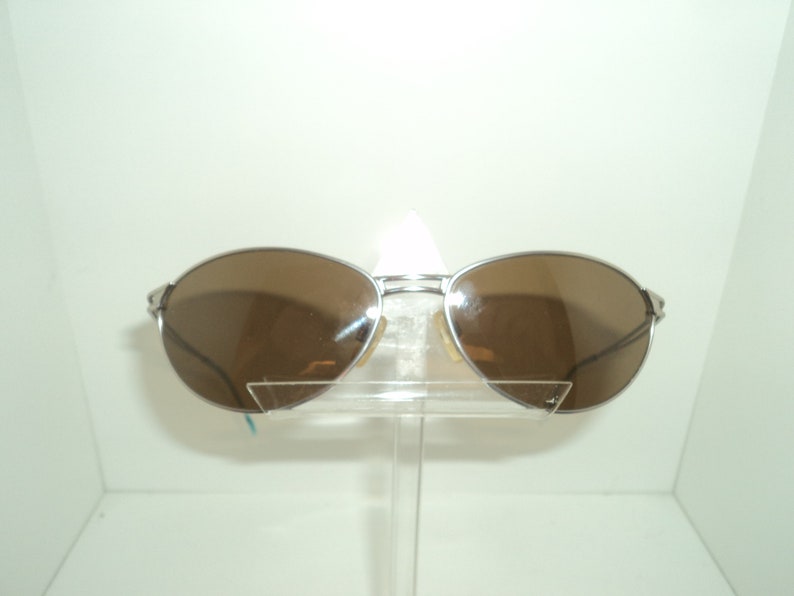 Puede incluir: Gafas de sol aviador plateadas con lentes marrones. Las gafas est&aacute;n en un expositor transparente.