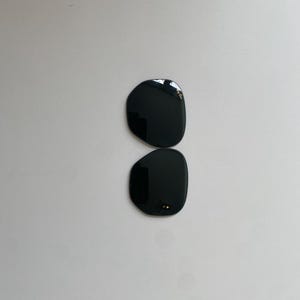 K&ouml;nnte beinhalten: Zwei separate, dunkel get&ouml;nte Ersatzgl&auml;ser f&uuml;r eine Sonnenbrille mit abgerundeter, unregelm&auml;&szlig;iger Form. Das obere Glas zeigt das wei&szlig;e Ray-Ban-Logo in der oberen Ecke, w&auml;hrend das untere Glas kleine Befestigungspunkte am Innenrand aufweist.