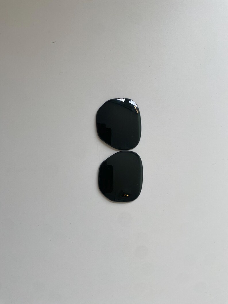 K&ouml;nnte beinhalten: Zwei separate, dunkel get&ouml;nte Ersatzgl&auml;ser f&uuml;r eine Sonnenbrille mit abgerundeter, unregelm&auml;&szlig;iger Form. Das obere Glas zeigt das wei&szlig;e Ray-Ban-Logo in der oberen Ecke, w&auml;hrend das untere Glas kleine Befestigungspunkte am Innenrand aufweist.