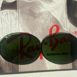 K&ouml;nnte beinhalten: Zwei ovale, dunkelgr&uuml;ne Brillengl&auml;ser aus Glas. Das linke Glas zeigt das wei&szlig;e Ray-Ban-Logo. Das rechte Glas tr&auml;gt den schwarzen Schriftzug Bausch & Lomb mit einem kleinen eingravierten Symbol. Die Gl&auml;ser liegen auf einer schwarz-wei&szlig;en Grafik.