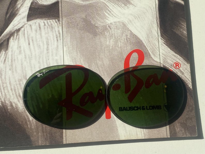 K&ouml;nnte beinhalten: Zwei ovale, dunkelgr&uuml;ne Brillengl&auml;ser aus Glas. Das linke Glas zeigt das wei&szlig;e Ray-Ban-Logo. Das rechte Glas tr&auml;gt den schwarzen Schriftzug Bausch & Lomb mit einem kleinen eingravierten Symbol. Die Gl&auml;ser liegen auf einer schwarz-wei&szlig;en Grafik.