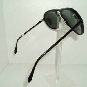 May include: Black aviator sunglasses with green lenses. The sunglasses are on a clear display stand.
