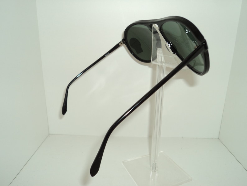 May include: Black aviator sunglasses with green lenses. The sunglasses are on a clear display stand.