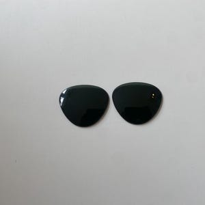 K&ouml;nnte beinhalten: Zwei einzelne Ersatzgl&auml;ser f&uuml;r eine Sonnenbrille mit einer glatten, durchgehend schwarzen Oberfl&auml;che. Jedes Glas hat eine abgerundete, dreieckige Form mit geschwungenen Kanten, die f&uuml;r das Einsetzen in einen Rahmen vorgesehen sind. Die Gl&auml;ser sind undurchsichtig.