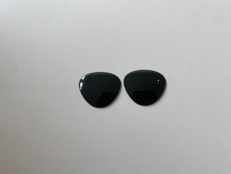 K&ouml;nnte beinhalten: Zwei einzelne Ersatzgl&auml;ser f&uuml;r eine Sonnenbrille mit einer glatten, durchgehend schwarzen Oberfl&auml;che. Jedes Glas hat eine abgerundete, dreieckige Form mit geschwungenen Kanten, die f&uuml;r das Einsetzen in einen Rahmen vorgesehen sind. Die Gl&auml;ser sind undurchsichtig.
