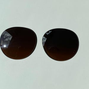 K&ouml;nnte beinhalten: Zwei runde, dunkelbraune Ersatzgl&auml;ser f&uuml;r Sonnenbrillen mit Farbverlauf. Das rechte Glas tr&auml;gt das wei&szlig;e Ray-Ban-Logo im oberen Bereich. Die Gl&auml;ser haben eine glatte, gew&ouml;lbte Oberfl&auml;che, auf der sich der Himmel in der dunklen T&ouml;nung spiegelt.