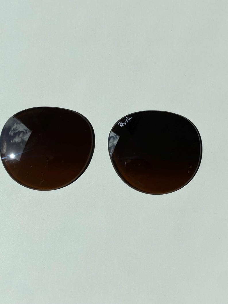 K&ouml;nnte beinhalten: Zwei runde, dunkelbraune Ersatzgl&auml;ser f&uuml;r Sonnenbrillen mit Farbverlauf. Das rechte Glas tr&auml;gt das wei&szlig;e Ray-Ban-Logo im oberen Bereich. Die Gl&auml;ser haben eine glatte, gew&ouml;lbte Oberfl&auml;che, auf der sich der Himmel in der dunklen T&ouml;nung spiegelt.