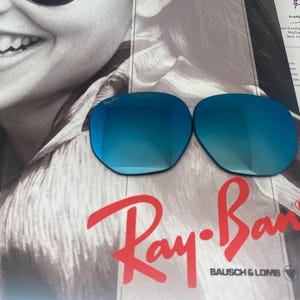 K&ouml;nnte beinhalten: Zwei Ersatzgl&auml;ser f&uuml;r eine Sonnenbrille mit reflektierender blauer Spiegelbeschichtung. Die Gl&auml;ser sind oval geformt und tragen das Ray-Ban-Logo, das in die obere Ecke des linken Glases ge&auml;tzt ist. Sie liegen auf einem Werbedruck mit dem Ray-Ban-Logo.