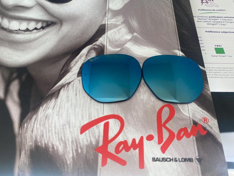 K&ouml;nnte beinhalten: Zwei Ersatzgl&auml;ser f&uuml;r eine Sonnenbrille mit reflektierender blauer Spiegelbeschichtung. Die Gl&auml;ser sind oval geformt und tragen das Ray-Ban-Logo, das in die obere Ecke des linken Glases ge&auml;tzt ist. Sie liegen auf einem Werbedruck mit dem Ray-Ban-Logo.