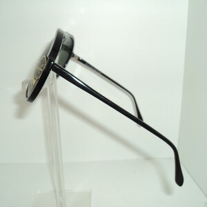 May include: Black aviator sunglasses with a gold logo on the side. The sunglasses are on a clear stand.