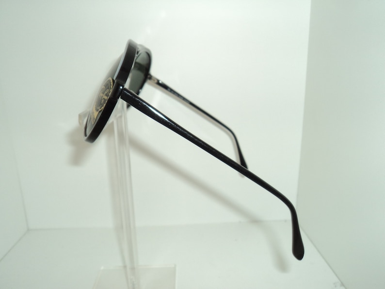 May include: Black aviator sunglasses with a gold logo on the side. The sunglasses are on a clear stand.
