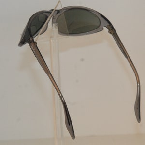 Pode incluir: &Oacute;culos de sol cinzentos com arma&ccedil;&atilde;o e lentes cinzas escuras. Os &oacute;culos est&atilde;o dobrados ao meio e as hastes est&atilde;o estendidas.