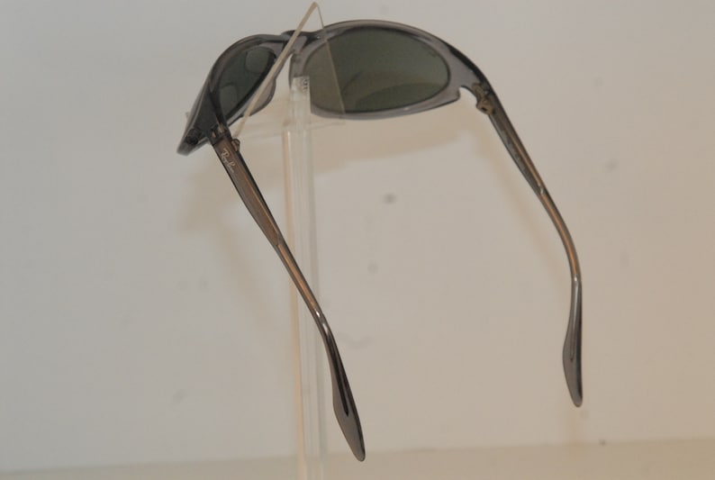 Pode incluir: &Oacute;culos de sol cinzentos com arma&ccedil;&atilde;o e lentes cinzas escuras. Os &oacute;culos est&atilde;o dobrados ao meio e as hastes est&atilde;o estendidas.