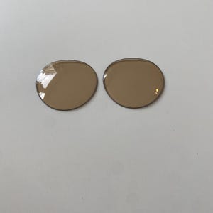 K&ouml;nnte beinhalten: Zwei kreisf&ouml;rmige, braun get&ouml;nte Ersatzgl&auml;ser f&uuml;r Brillen mit einer glatten, polierten Oberfl&auml;che. Diese Gl&auml;ser sind f&uuml;r Brillenfassungen konzipiert und weisen eine durchgehend gleichm&auml;&szlig;ige hellbraune Farbe mit sichtbaren Lichtreflexen auf.