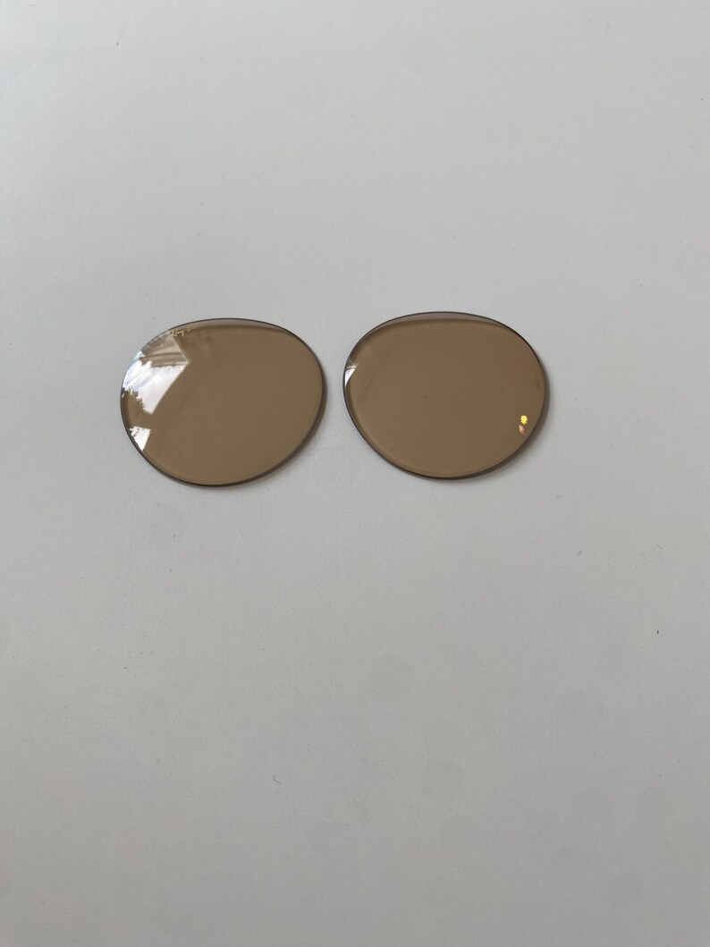 K&ouml;nnte beinhalten: Zwei kreisf&ouml;rmige, braun get&ouml;nte Ersatzgl&auml;ser f&uuml;r Brillen mit einer glatten, polierten Oberfl&auml;che. Diese Gl&auml;ser sind f&uuml;r Brillenfassungen konzipiert und weisen eine durchgehend gleichm&auml;&szlig;ige hellbraune Farbe mit sichtbaren Lichtreflexen auf.