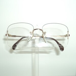 May include: A pair of rimless eyeglasses with a silver frame and brown temples. The glasses are on a clear plastic stand.