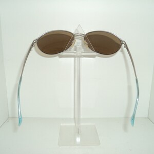 Puede incluir: Gafas de sol plateadas con lentes marrones, expuestas en un soporte de pl&aacute;stico transparente. Las patillas de las gafas son de color azul claro.