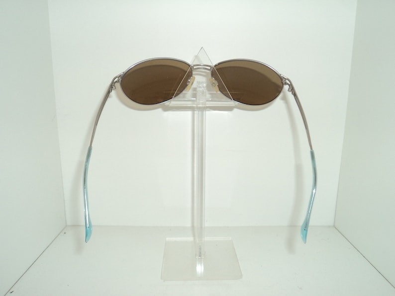 Puede incluir: Gafas de sol plateadas con lentes marrones, expuestas en un soporte de pl&aacute;stico transparente. Las patillas de las gafas son de color azul claro.