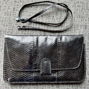 Puede incluir: Bolso de mano negro con estampado de piel de reptil, cierre de solapa y correa de hombro desmontable. El bolso es rectangular y tiene un acabado brillante. La correa es fina y negra.