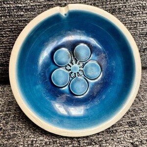 May include: A round, ceramic ashtray with a vibrant blue interior and a cream-colored rim. The center features a raised floral design in a lighter shade of blue. The ashtray has a small notch for resting cigarettes.