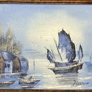 Pode incluir: Pintura a óleo de uma cena costeira com barcos e edifícios. A obra de arte apresenta uma paleta de cores azul e branca, com uma moldura de madeira. A pintura retrata vários veleiros com velas azul-escuras na água e edifícios na costa.