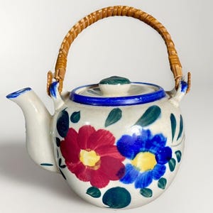 May include: A ceramic teapot with a woven handle. The teapot is white with a blue rim and lid, and is decorated with red and blue floral designs and green leaves. The handle is made of woven brown material.