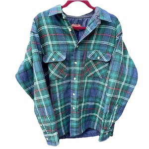 Puede incluir: Una camisa de franela a cuadros azules, verdes y rojos con dos bolsillos en el pecho y botones. La camisa está colgada de una percha roja. La camisa tiene un forro acolchado.