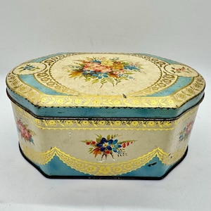 May include: An antique, octagonal tin box with a floral design. The lid and sides feature a cream-colored background with a blue border and gold accents. The lid is adorned with a bouquet of colorful flowers.