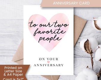 Puede incluir: Una tarjeta blanca con un corazón rosa de acuarela con el texto "to our two favorite people" y "ON YOUR ANNIVERSARY". La tarjeta mide 5x7 pulgadas cuando está doblada.