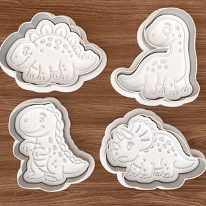 Pode incluir: Quatro cortadores de biscoitos de plástico branco em forma de dinossauro. Os cortadores incluem um Estegossauro, um Brontossauro, um Tiranossauro Rex e um Triceratops. Os cortadores estão em uma superfície de madeira.
