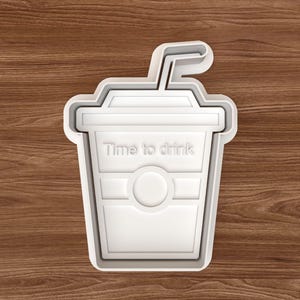 May include: A white plastic cookie cutter shaped like a takeaway drink cup with a straw. The words "Time to drink" are embossed on the front. The cutter is on a wooden surface.