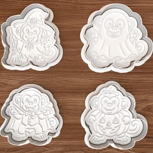 May include: A set of four white plastic cookie cutters featuring monkey characters in Halloween costumes. The designs include a monkey as a witch with a broom, a monkey dressed as a ghost, a monkey as a vampire, and a monkey inside a pumpkin.