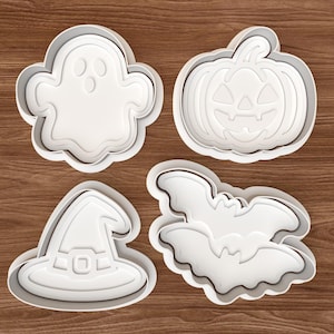 Puede incluir: Cuatro cortadores de galletas blancos con temática de Halloween: un fantasma, una calabaza, un sombrero de bruja y un murciélago. Cada cortador tiene un diseño en capas, creando un efecto 3D. Los cortadores están dispuestos sobre una superficie de madera.